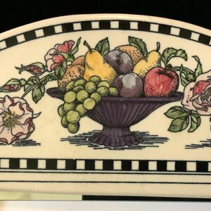 May include: A white ceramic plate with a black and white checkered border. The center of the plate features a hand-painted design of a fruit bowl filled with pears, plums, grapes, and an apple. The bowl is surrounded by roses and leaves.