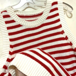May include: A sleeveless, striped sweater in red and white. The sweater features horizontal stripes and a ribbed neckline and armholes. The garment is made of a knitted material and is hanging on a clear plastic hanger.