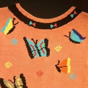 K&ouml;nnte beinhalten: Ein rosa gestrickter Pullover mit schwarzem Besatz am Ausschnitt und bunten Schmetterlingsmotiven.