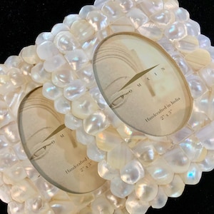 May include: Two oval-shaped picture frames made of white mother of pearl shell tiles. The frames are 2 inches by 3 inches and are handcrafted in India. The frames are empty and ready for photos.