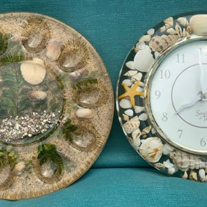 May include: Two decorative items: a round resin piece with sand, shells, and greenery, and a clock with a silver frame and seashells. The clock face is white with black numbers and hands. The clock reads "Sands of Time".