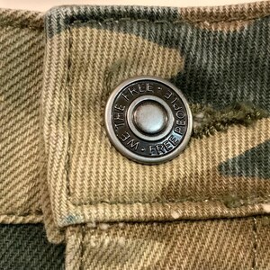 May include: Close-up of a button on camouflage-patterned fabric. The silver button is embossed with the words "WE FREE PEOPLE FREE" around a central circle. The fabric is a mix of green, brown, and tan.