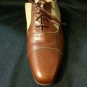 May include: Brown leather Oxford shoes with a beige canvas panel and laces. The shoes have a wingtip design and a polished finish.