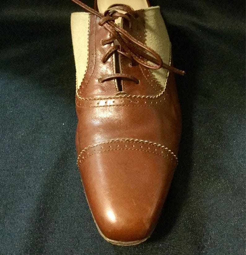 May include: Brown leather Oxford shoes with a beige canvas panel and laces. The shoes have a wingtip design and a polished finish.