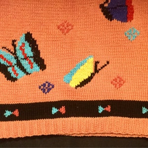 K&ouml;nnte beinhalten: Ein rosa gestrickter Pullover mit schwarzem Saum und einem bunten Schmetterlingsmuster. Der Pullover hat verschiedene farbige Schmetterlinge und geometrische Formen.