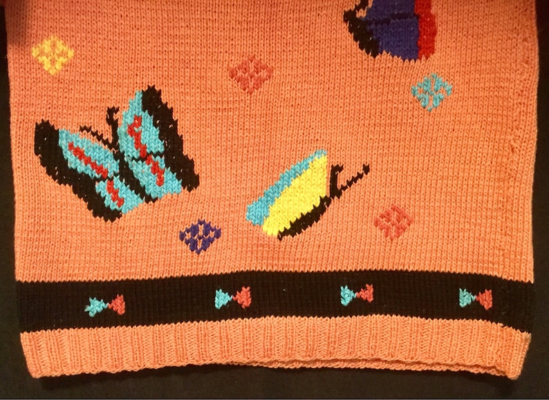 K&ouml;nnte beinhalten: Ein rosa gestrickter Pullover mit schwarzem Saum und einem bunten Schmetterlingsmuster. Der Pullover hat verschiedene farbige Schmetterlinge und geometrische Formen.