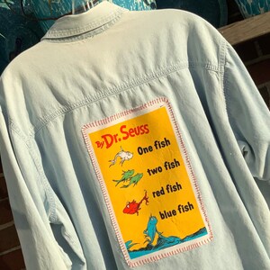 May include: Light blue denim shirt with a colorful patch featuring illustrations of fish and the text "One fish, two fish, red fish, blue fish" by Dr. Seuss. The patch is rectangular with a red stitched border.