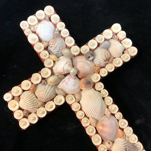 May include: A decorative cross crafted from a red base, adorned with various seashells and small, white faux pearls. The shells are in shades of beige, brown, and white, creating a textured surface. The cross is a unique, handcrafted item.