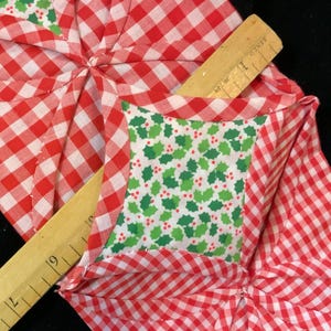 May include: A red and white gingham fabric with a green and white holly leaf print fabric sewn into the center. The fabric is folded into a star shape.