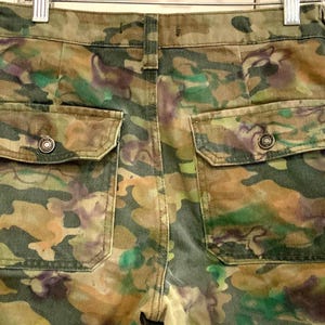 May include: Camouflage-patterned pants with two back pockets. The pants feature a mix of green, brown, and purple hues, creating a unique camouflage design. The pockets have button closures. The pants are hanging on a metal hanger.