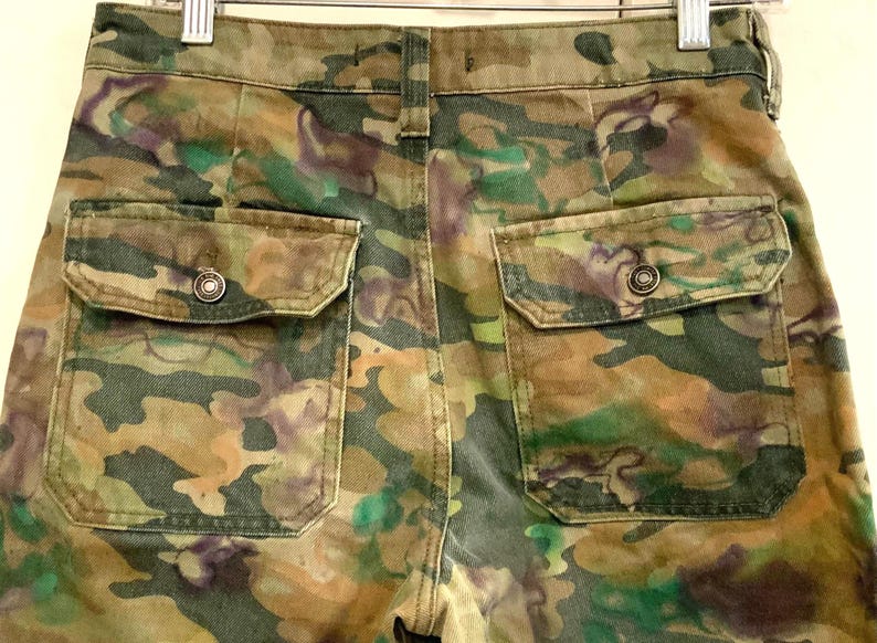 May include: Camouflage-patterned pants with two back pockets. The pants feature a mix of green, brown, and purple hues, creating a unique camouflage design. The pockets have button closures. The pants are hanging on a metal hanger.