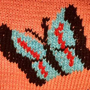 K&ouml;nnte beinhalten: Ein gestrickter Stoff mit einem Schmetterlingsmuster in Schwarz, Blau und Rot. Der Schmetterling befindet sich mittig auf einem pfirsichfarbenen Hintergrund.