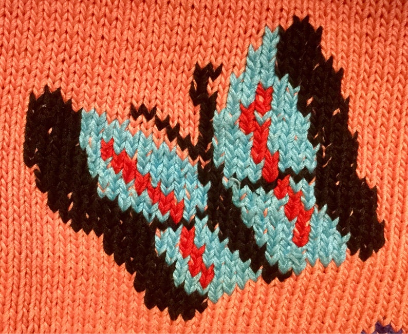 K&ouml;nnte beinhalten: Ein gestrickter Stoff mit einem Schmetterlingsmuster in Schwarz, Blau und Rot. Der Schmetterling befindet sich mittig auf einem pfirsichfarbenen Hintergrund.