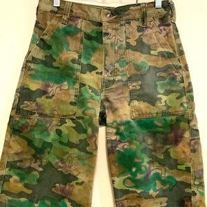 May include: Camouflage-patterned pants in shades of green, brown, and purple. The pants feature two front pockets and a button closure. The fabric has a faded appearance, giving it a vintage look. The pants are hanging on a metal hanger.