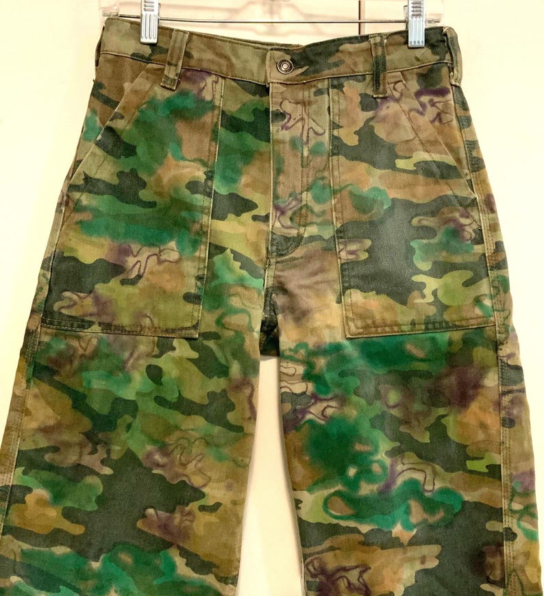 May include: Camouflage-patterned pants in shades of green, brown, and purple. The pants feature two front pockets and a button closure. The fabric has a faded appearance, giving it a vintage look. The pants are hanging on a metal hanger.