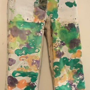 May include: White denim pants with a camouflage waistband and fly. The pants are decorated with colorful paint splatters in shades of green, orange, and purple. The pants are hanging on a metal hanger.