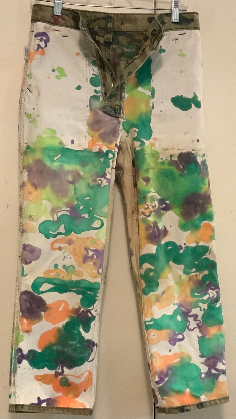 May include: White denim pants with a camouflage waistband and fly. The pants are decorated with colorful paint splatters in shades of green, orange, and purple. The pants are hanging on a metal hanger.