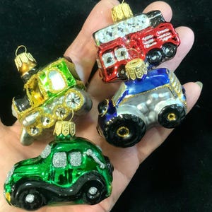 May include: Four miniature glass vehicle ornaments. The ornaments include a green car, a blue tractor, a red double-decker bus, and a green train. Each ornament has gold accents and a loop for hanging.