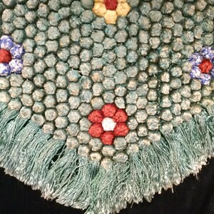 May include: A teal blue rug with a fringe border and a pattern of colorful flowers. The flowers are made of fabric and are sewn onto the rug.