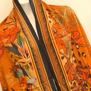 May include: A long orange scarf with a black and gold striped border. The scarf features a floral print with orange, yellow, and green flowers.