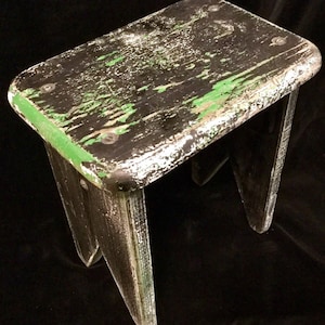 May include: A small wooden stool with a black and green paint finish. The paint is chipped and worn, giving the stool a rustic look.