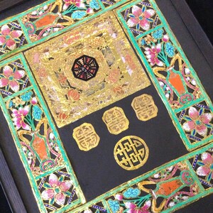 May include: A framed artwork with a black background and intricate gold and green paper cutouts. The artwork features a central square with a gold background and a black and gold design. The square is surrounded by four smaller gold squares with Chinese characters. The artwork is framed in a black frame.