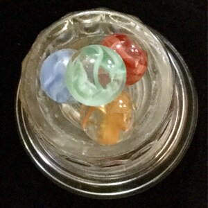 May include: Four glass marbles in a clear glass jar. The marbles are blue, green, orange, and red. The marbles have swirls of color.