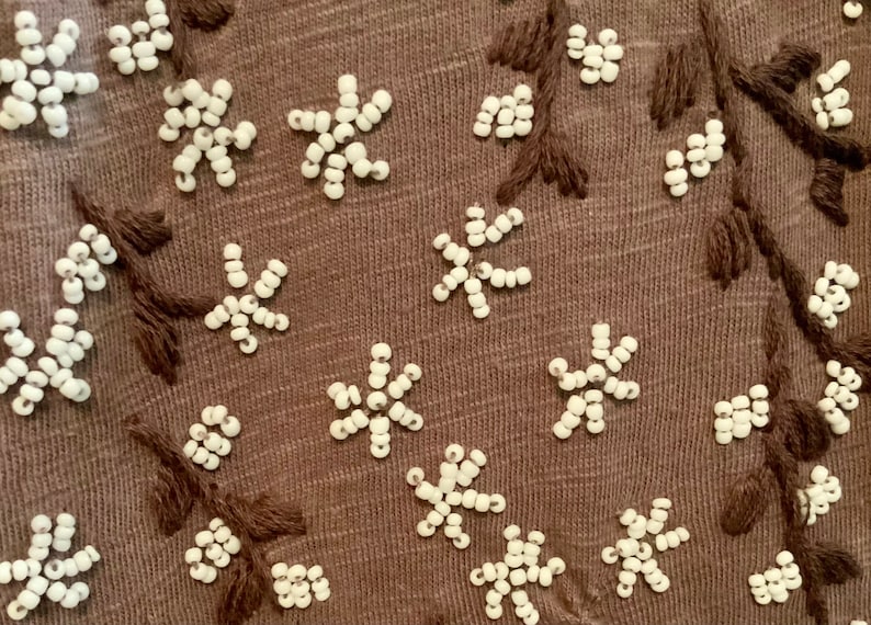 May include: Close-up of a brown fabric with white bead embroidery in the shape of flowers and branches.