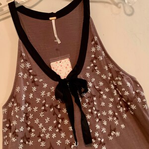 May include: A brown sleeveless top with a black trim around the neckline and a black bow at the neck. The top is decorated with white embroidered flowers.