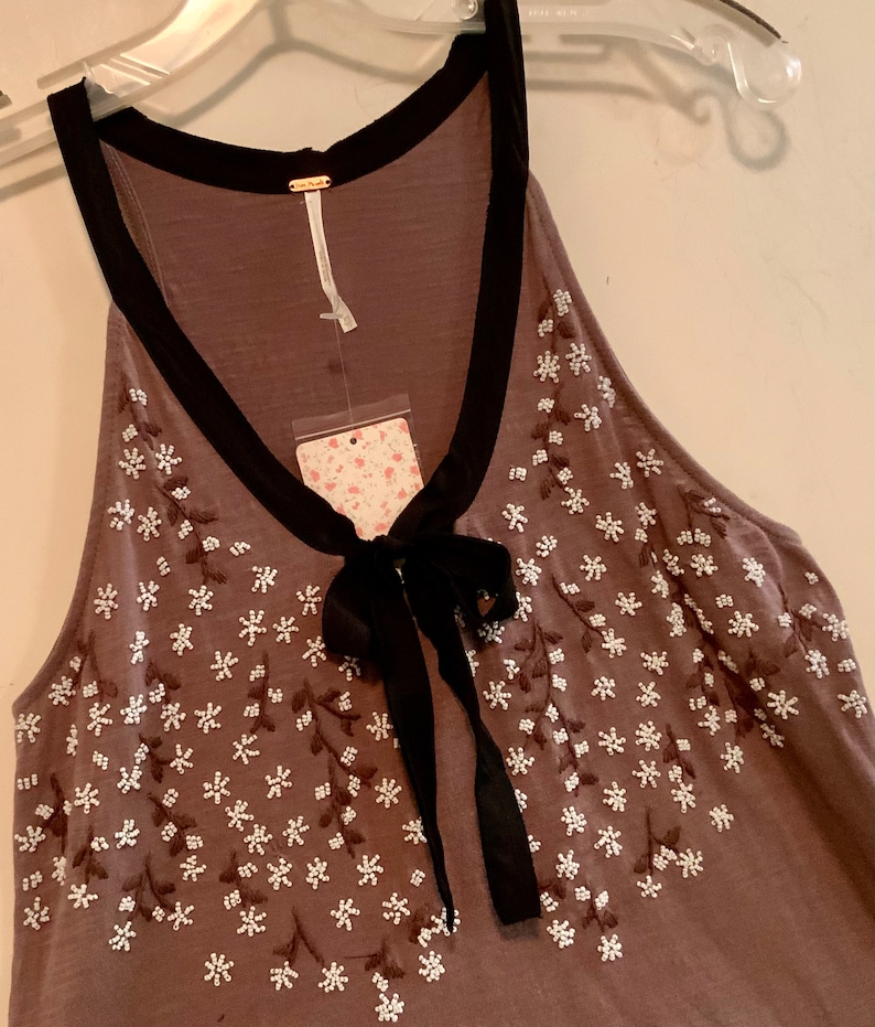 May include: A brown sleeveless top with a black trim around the neckline and a black bow at the neck. The top is decorated with white embroidered flowers.