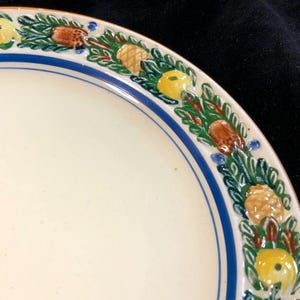 May include: Cream-colored ceramic plate with a blue border and a decorative rim featuring green leaves, yellow fruit, and brown pinecones. The plate is partially visible, showcasing a portion of its design.
