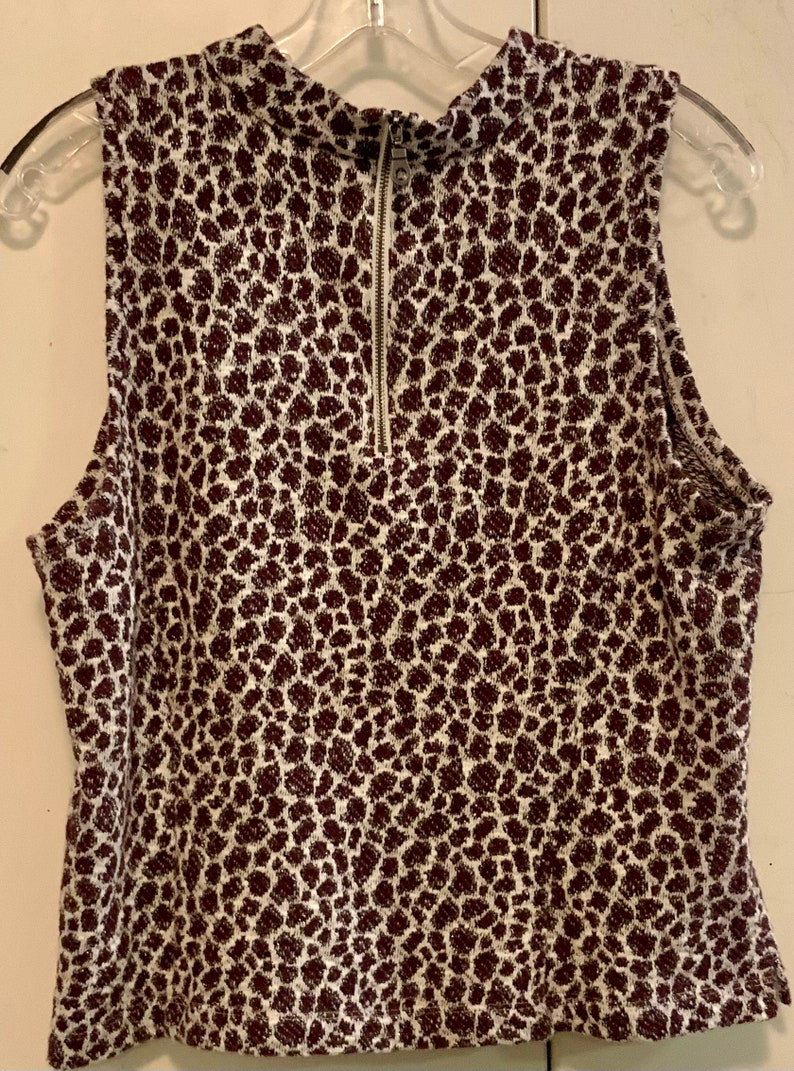 Puede incluir: Una blusa sin mangas blanca con estampado de leopardo marr&oacute;n. La blusa tiene cuello alto y una cremallera en la parte delantera.