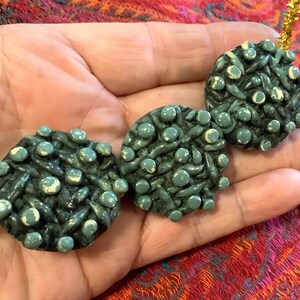 May include: A handmade necklace featuring three dark teal, textured, circular beads. Each bead is adorned with small, light teal spheres and white accents, strung on a gold-colored cord. The beads have a woven appearance.