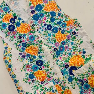 May include: A white sleeveless vest with a floral pattern in shades of blue, yellow, pink, and green. The vest has a V-neck and a button closure.