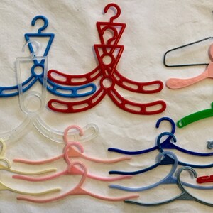 May include: A collection of 14 plastic hangers in various colors, including clear, pink, red, blue, green, and white. Some hangers are designed with multiple hooks, while others have a single hook. The hangers are arranged on a white surface.