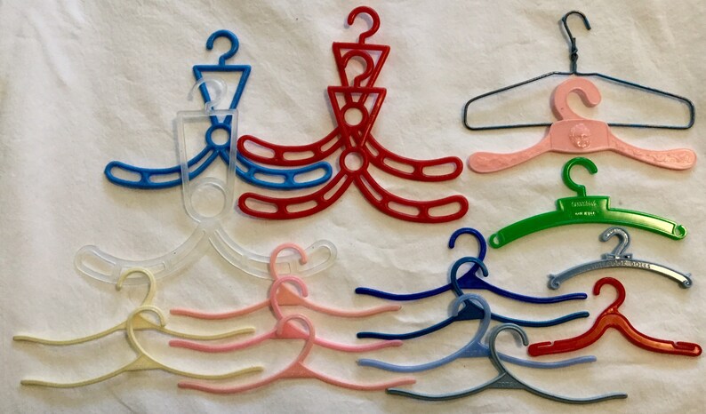 May include: A collection of 14 plastic hangers in various colors, including clear, pink, red, blue, green, and white. Some hangers are designed with multiple hooks, while others have a single hook. The hangers are arranged on a white surface.