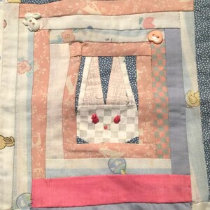 May include: A handmade quilt with a central bunny design. The quilt features a patchwork design with pink, blue, and white fabrics. A small bunny button and embroidered details add charm. The quilt is a cozy, handcrafted item.