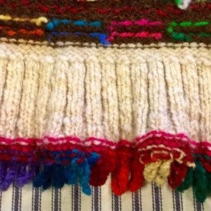 K&ouml;nnte beinhalten: Ein gestricktes Kleidungsst&uuml;ck mit einem wei&szlig;en Fransenbesatz und einem bunten bestickten Rand. Die Fransen sind aus wei&szlig;em Garn und der Rand aus rotem, rosa, blauem, gr&uuml;nem und gelbem Garn.