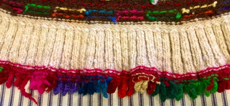 K&ouml;nnte beinhalten: Ein gestricktes Kleidungsst&uuml;ck mit einem wei&szlig;en Fransenbesatz und einem bunten bestickten Rand. Die Fransen sind aus wei&szlig;em Garn und der Rand aus rotem, rosa, blauem, gr&uuml;nem und gelbem Garn.