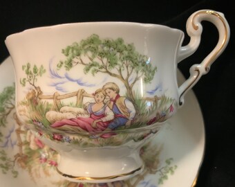May include: A white teacup and saucer with a gold trim. The teacup features a painted scene of a couple in a pastoral setting with a tree, fence, and sheep. The saucer has a similar design.