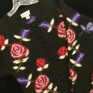 May include: Black knitted cardigan with a floral pattern featuring red, pink, and purple roses. The cardigan has a button closure and the label "Rosanna" is visible.