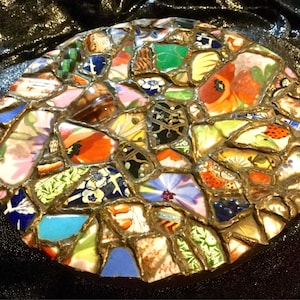 May include: A round mosaic made from colorful ceramic shards. The mosaic features a variety of patterns and colors, including floral designs, geometric shapes, and abstract patterns. The pieces are set in a gold-colored grout, creating a decorative piece.