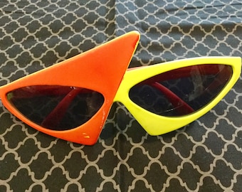 Könnte beinhalten: Eine Sonnenbrille im Retro-Stil mit einem auffälligen, kantigen Design. Ein Rahmen ist leuchtend orange, der andere gelb. Beide haben dunkle Gläser und rote Bügel. Die Sonnenbrille liegt auf einem gemusterten Stoff.