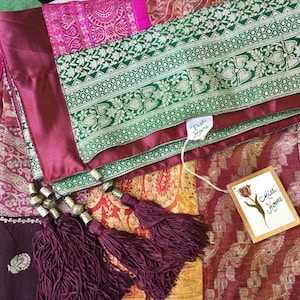May include: A multi-colored fabric with a green and gold patterned border. The fabric has a burgundy trim and is decorated with tassels and metal beads. The fabric has two tags attached to it, one that says "Tuli Home" and another that says "Mili Home".