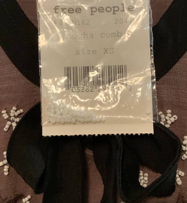 May include: A close-up of a white tag with black text that reads "free people" and "mocha comb" with a barcode and the text "size XS". The tag is attached to a brown garment with white beads and a black ribbon.