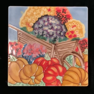 May include: Square ceramic tile featuring a fall harvest scene. The image depicts a brown wheelbarrow surrounded by pumpkins in shades of orange and red. Colorful flowers in the background add to the autumn theme. The tile has a glossy finish.