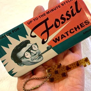 May include: A vintage-style tin box for Fossil watches. The box is green, red, and white with the text "Watch for... Up-to-the-minute style Fossil Watches Genuine". The box features a cartoon man wearing glasses with clocks on the lenses and smoking a pipe.