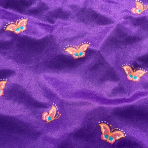 Op de afbeelding: Paarse stof met een herhalend patroon van kleurrijke vlinders. De vlinders hebben oranje, roze en blauwe accenten.