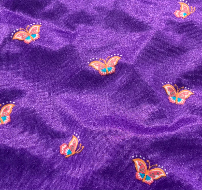 Op de afbeelding: Paarse stof met een herhalend patroon van kleurrijke vlinders. De vlinders hebben oranje, roze en blauwe accenten.