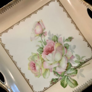 May include: A square porcelain serving tray with a gold trim and a floral design featuring pink and white roses. The tray has two handles on the sides.